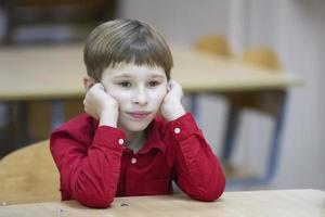 Child primary school student. A boy in a red shirt at the table. photo