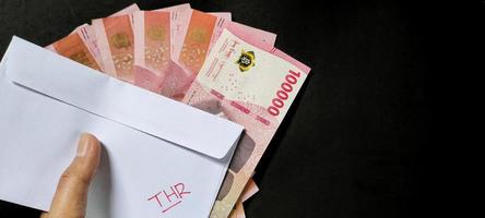 Man holding a white envelope written of THR and new Indonesian banknotes, usually Tunjangan Hari Raya or called THR are given to employees ahead of Eid. Isolated on black background and top view photo