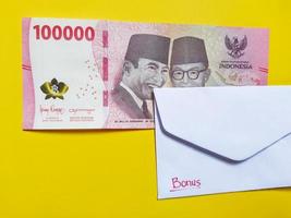A white envelope written of bonus and new Indonesian banknotes, usually Tunjangan Hari Raya or called THR are given to employees ahead of Eid. Isolated on yellow background and top view photo