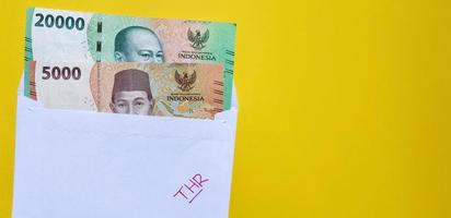 A white envelope written of THR and new Indonesian banknotes, usually Tunjangan Hari Raya or called THR are given to employees ahead of Eid. Isolated on yellow background and negative space photo