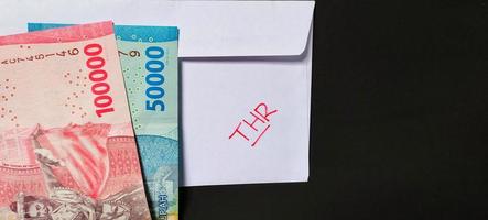 A white envelope written of THR and new Indonesian banknotes, usually Tunjangan Hari Raya or called THR are given to employees ahead of Eid. Isolated on a black background photo