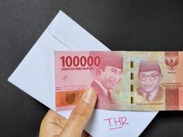 Man holding a white envelope written of THR and new Rp.100,000 Indonesian banknotes, usually Tunjangan Hari Raya or called THR are given to employees ahead of Eid. Top view photo