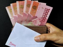 Man holding a white envelope written bonus and new Indonesian banknotes. Usually the Holiday Allowance or known as THR is given to employees ahead of Eid. Top view photo