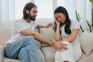 A sad Asian woman talks to a man in tears at home sitting on the couch. Young couple of different nationalities and conflict of interest in a couple photo