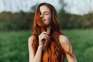 A young woman smiles and looks into the camera with her long, wavy, shiny red hair in a park with green grass in the summer sunset photo