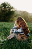 un mujer se sienta en el verde césped en un parque con su espalda a el cámara y relaja en naturaleza en el verano puesta de sol noche ligero foto