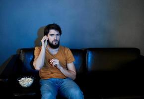 un hombre se sienta en un sofá adentro y relojes televisión frente ver foto