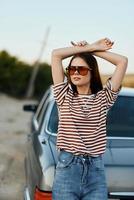 elegante mujer en a rayas camiseta Gafas de sol y pantalones cerca gris coche en naturaleza foto