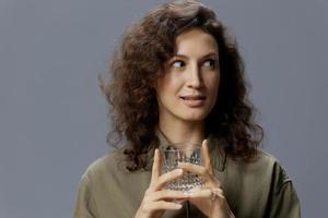 Dreaming curly beautiful woman in casual khaki green shirt hold glass of water thinking looks aside posing isolated on over gray blue background. Healthy lifestyle. Water is Life concept. Copy space photo