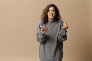 Excited happy curly beautiful lady in gray casual sweater get cool news from message posing isolated on over beige pastel background. Social media, network, distance communication concept. Copy space photo
