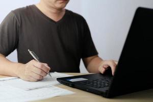 hombre utilizando ordenador portátil computadora y tomando notas en planificación. cuales es pago en línea, se sienta en el silla en el vivo habitación a hogar en un ordenador portátil. el concepto de Finanzas y en línea compras. foto