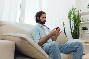 A freelance man in a white T-shirt, blue jeans, and shirt sits on the couch with his phone at home on his day off and plays games photo