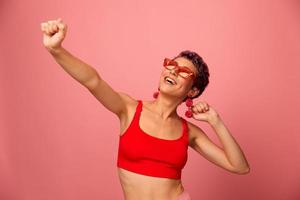 A young athletic woman with a short haircut and purple hair in a red top in sunglasses with an athletic figure smiles with her arms outstretched in different directions dancing on a pink background photo