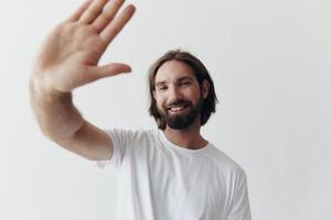 Happy adult man with a beard smiles and pulls the cook into the camera listening to music in headphones in a distressed t-shirt on a white isolated background photo