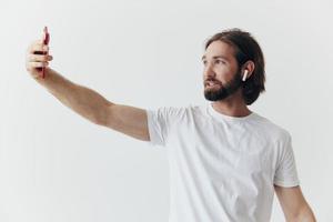 A male blogger videotaping himself on his phone and chatting with people online with a smile in a white T-shirt against a white wall photo