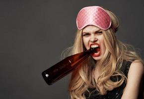 enojado mujer con un botella de cerveza en su mano con brillante maquillaje y un rosado dormir máscara foto