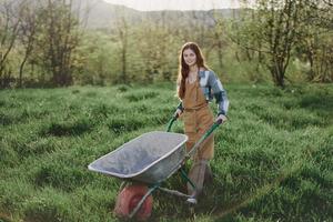 A happy woman with a cart works in her country home in the countryside against a backdrop of green grass and sunset sun photo