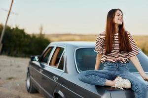 un joven mujer se sienta en el maletero de un coche y descansa después un difícil la carretera y admira naturaleza con un hermosa vista. parada es además parte de el viaje foto