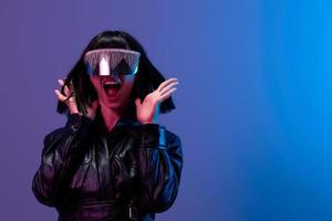 Crazy pretty awesome brunet woman in leather jacket trendy specular sunglasses scream spread hands posing isolated in blue violet color light background. Neon party Cyberpunk concept. Copy space photo