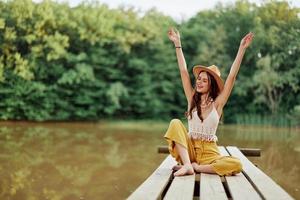 Hippie eco-activist woman traveler sits on a bridge by a lake with her arms outstretched with a hat and smiling sincerely photo