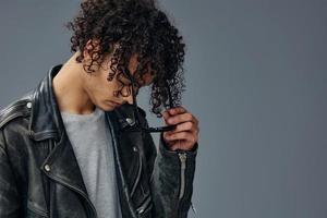 Side of view portrait of stylish tanned curly man leather jacket wear glasses posing isolated on over gray background. Cool fashion offer. Huge Seasonal Sale New Collection concept. Copy space for ad photo