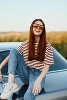 A fashionable woman smiles sweetly in stylish sunglasses, a striped t-shirt and jeans, sits on the trunk of a car and looks at the beautiful nature of autumn. Travel lifestyle photo