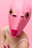 A woman wearing a silicone Halloween mask in the shape of a pink fish with big yellow eyes looks at the camera against a pink background photo