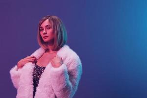 Beautiful adorable blonde lady wear touch fluffy fur coat sparkly dress look at camera posing isolated in blue violet pink color light studio background. Neon party Fashion concept. Copy space Banner photo