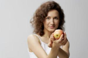 sonriente encantador linda Rizado hermosa mujer en básico blanco camiseta tira manzana disfrutar sano comida posando aislado en terminado blanco antecedentes. natural Respetuoso del medio ambiente productos concepto. Copiar espacio foto