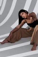 A beautiful lady in beige pants and a black top sits on the floor and a falling shadow in a bright room photo