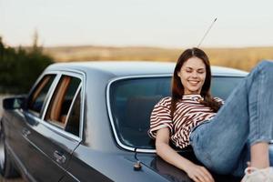 The woman driver stopped on the road and lay down on the car to rest and look at the beautiful landscape in a striped T-shirt and jeans. Complicated journey to nature photo