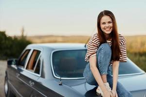 A young woman sits on the trunk of a car, laughs and rests after a difficult road and admires nature with a beautiful view. Stopping is also part of the journey photo