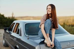 un joven mujer se sienta en el maletero de un coche y descansa después un difícil la carretera y admira naturaleza con un hermosa vista. parada es además parte de el viaje foto