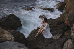 bonito mujer con mojado pelo en blanco vestir se sienta en olas piedras foto