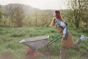 A young woman rolls a garden cart with soil for planting in her green nature garden and smiles photo