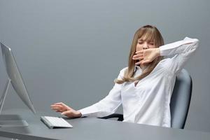 Sleepy tired pretty blonde businesswoman worker freelancer using desktop computer yawns sitting at workplace in gray modern office. Remote Job, Technology And Career Profession Concept. Copy space photo