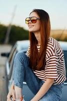 A fashionable woman smiles sweetly in stylish sunglasses, a striped t-shirt and jeans, sits on the trunk of a car and looks at the beautiful nature of autumn. Travel lifestyle photo