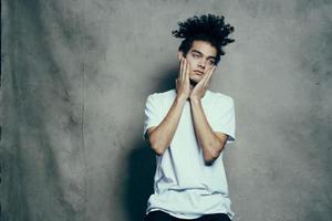 Cheerful guy with curly hair gestures with his hands near his face on a gray fabric background in a white t-shirt photo