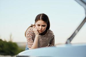 A woman with a wrench looks sadly and sadly under the open hood of her car and can't fix it from a roadside breakdown while traveling alone photo