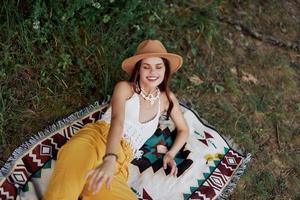 un mujer en Respetuoso del medio ambiente ropa en un hippie Mira es acostado en un de colores tartán sonriente y mirando a un otoño puesta de sol en naturaleza en el parque. estilo de vida en un viaje de armonía con el mundo foto