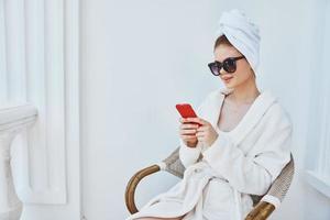 un mujer en un blanco túnica se sienta en un silla con un rojo teléfono en oscuro lentes aquí o en el Mañana foto