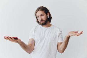 un hombre con un barba blogger en un blanco camiseta con un teléfono y inalámbrico auriculares mira dentro el teléfono y se extiende su manos a el lado en un blanco antecedentes aislado Copiar espacio foto