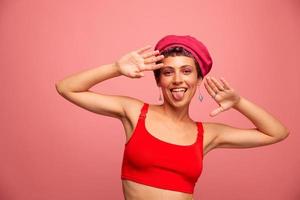 joven atlético mujer con un corto Corte de pelo y púrpura pelo en un rojo parte superior y un rosado sombrero con un atlético figura sonrisas y muecas mirando a el cámara en un rosado antecedentes