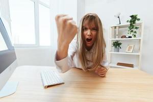 Angry evil blonde businesswoman screaming to subordinates shaking fist at camera sitting at workplace in modern office. Irritated director work on computer online in financial corporation. Wide angle photo