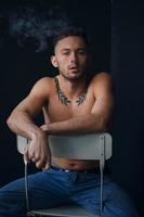 Modelling snapshots. Pensive frustrated tanned attractive handsome naked man sit on chair smoking looks at camera posing isolated in black studio background. Fashion offer. Copy space for ad. Closeup photo