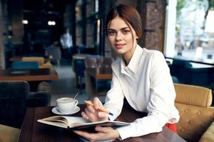 a woman in a shirt with a notebook and a pen in her hand sits at a table in a restaurant on an upholstered chair photo