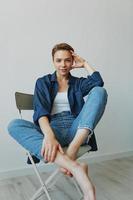 A young woman sitting in a chair at home smiling with teeth with a short haircut in jeans and a denim shirt on a white background. Girl natural poses with no filters photo