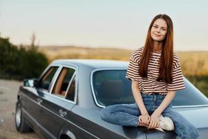 un joven mujer se sienta en el maletero de un coche y descansa después un difícil la carretera y admira naturaleza con un hermosa vista. parada es además parte de el viaje foto
