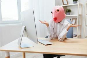 Pensive crazy businesswoman worker freelancer in pink fish mask work on computer online pondering or making decision thinking of problem solution spread hands in light modern office. Copy space photo