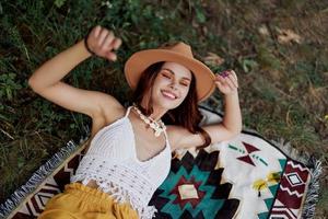 un mujer en Respetuoso del medio ambiente ropa en un hippie Mira es acostado en un de colores tartán sonriente y mirando a un otoño puesta de sol en naturaleza en el parque. estilo de vida en un viaje de armonía con el mundo foto